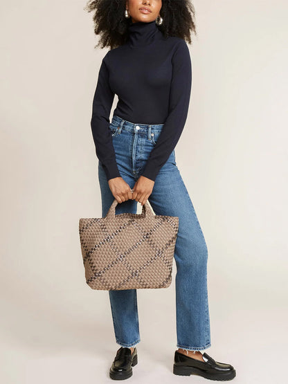 A woman in a black turtleneck and blue jeans holds a Naghedi St. Barths Medium Tote in Plaid Mojave while standing in a neutral pose.