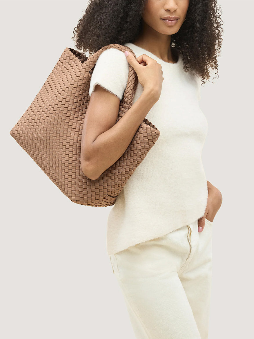 A person wearing a white top carries a large tan neoprene Naghedi St. Barths Medium Tote in Seychelles over their shoulder as an everyday carryall.