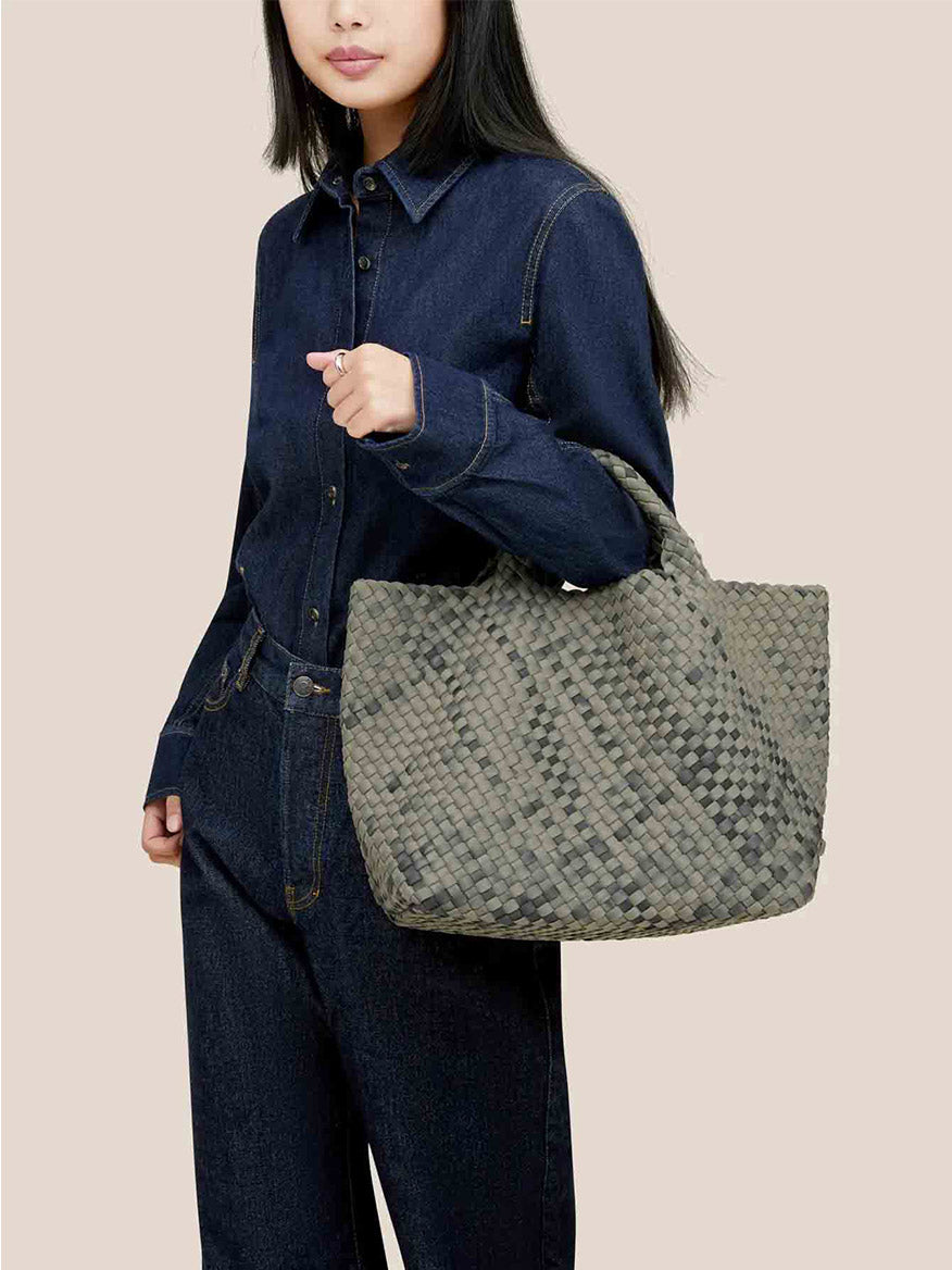 Dressed in denim, a person carries the sophisticated Naghedi St. Barths Medium Tote in Watercolor Neptune, featuring an elegant handwoven neoprene design.