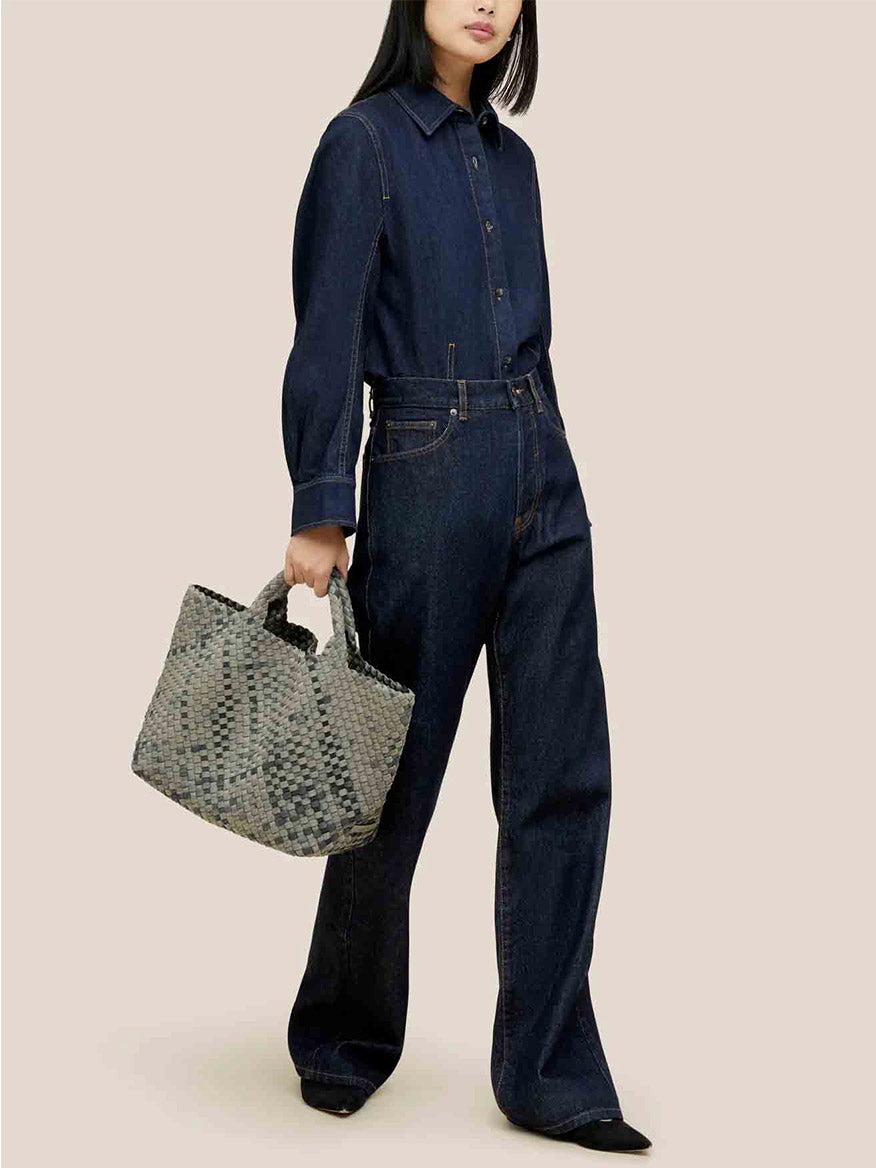 A person dressed in a denim shirt and pants holds the Naghedi St. Barths Medium Tote in Watercolor Neptune against a beige background.