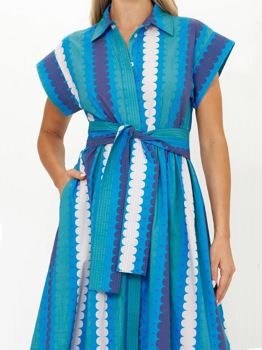 A person is wearing the Oliphant Belted Shirt Dress in Meko Blue, a blue and teal, midi-length shirt dress with white and dark purple stripes. Made from cotton poplin, it features a collar, short sleeves, a matching belt, and pockets where one hand rests casually.