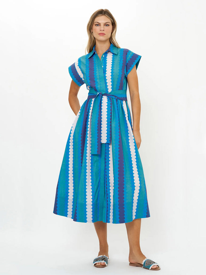 A woman wears the Oliphant Belted Shirt Dress in Meko Blue, a blue and white patterned cotton poplin piece with short sleeves and a belt, reaching midi length. She finishes her look with sandals against a plain white background.