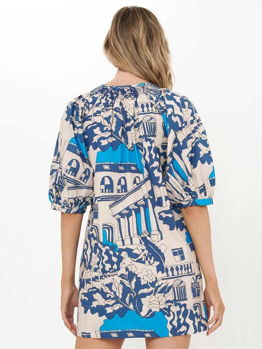Woman stands with her back to the camera, wearing the Oliphant Embroidered Button Mini Dress in Roma Blue, a cotton poplin mini shift dress featuring a blue and cream architectural pattern and short puff sleeves.