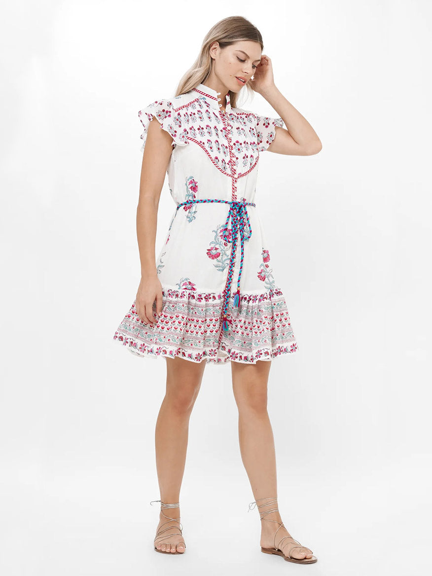 A woman stands against a plain white background wearing the Oliphant High Neck Button Mini Dress in Napa Red, featuring pink floral patterns, paired with a blue braided waist belt and tan strappy sandals.