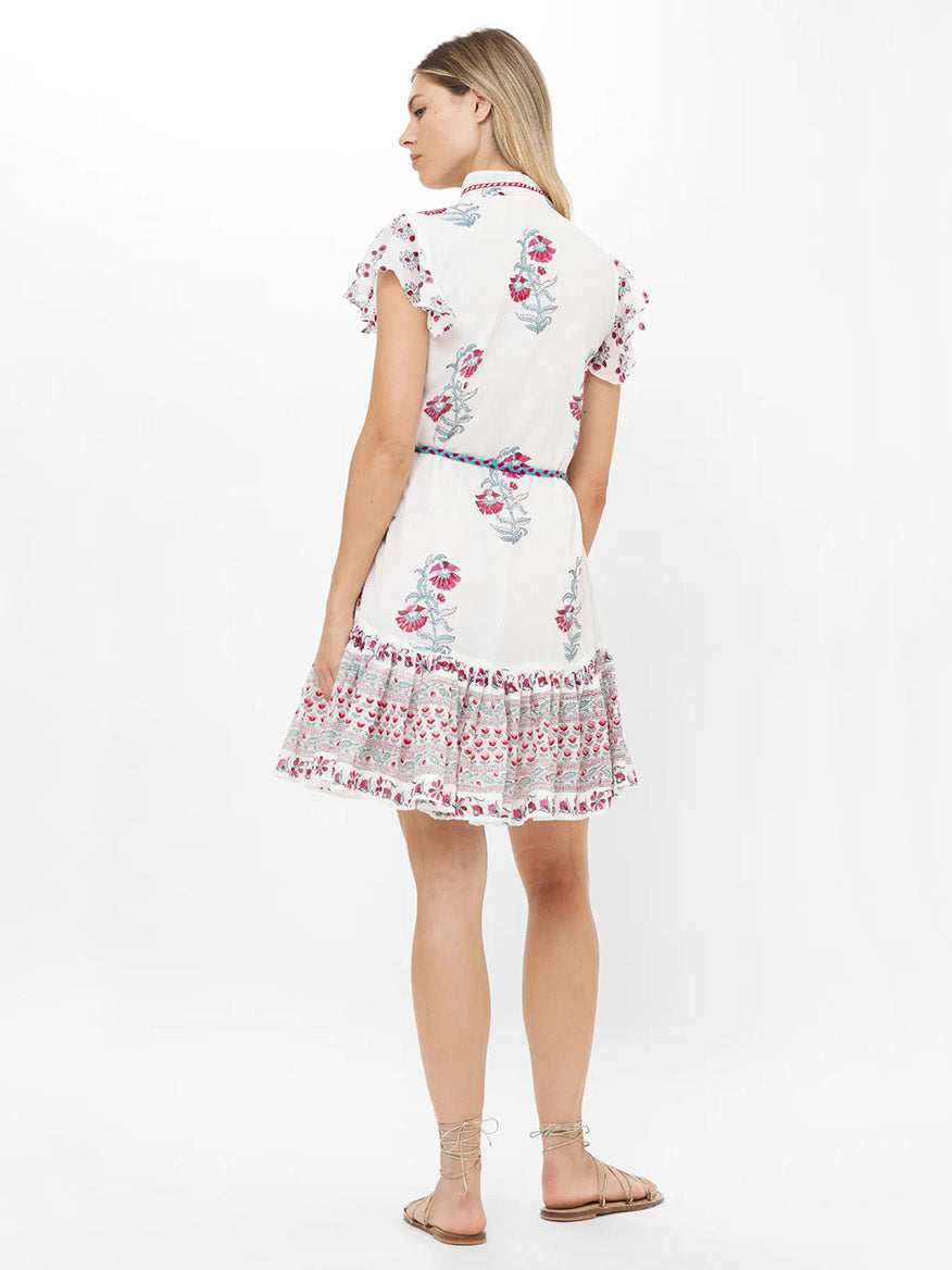 A woman stands facing away, wearing the Oliphant High Neck Button Mini Dress in Napa Red, a white cotton voile dress with pink floral patterns, ruffled hem, and short sleeves, paired with flat sandals.