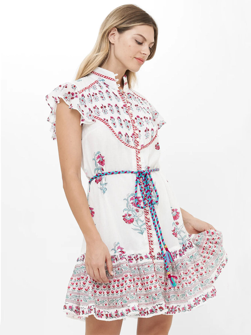 Woman wearing the Oliphant High Neck Button Mini Dress in Napa Red, a white cotton voile dress with short sleeves, floral pattern, ruffled shoulders, and a blue and red braided belt, against a plain white background.