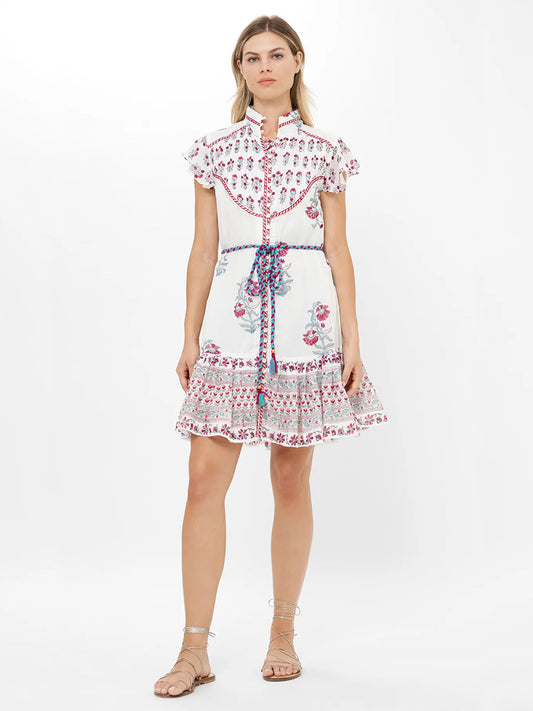 A woman stands against a plain white background wearing the Oliphant High Neck Button Mini Dress in Napa Red with a mandarin collar, ruffled sleeves, pink floral patterns, blue and pink braided belt, and flat sandals.