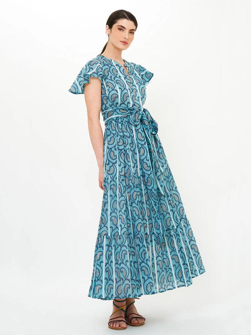 A woman stands wearing the Oliphant V-Neck Maxi Dress in Goa Blue, featuring hand block prints, flutter sleeves, a tied waist, and brown sandals against a plain light background.