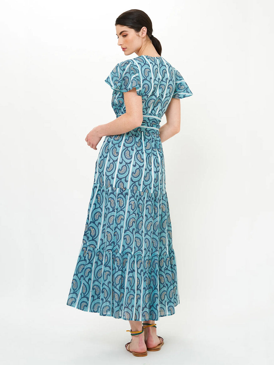 A woman, facing slightly away, wears the Oliphant V-Neck Maxi Dress in Goa Blue with flutter sleeves and sandals against a plain light background.