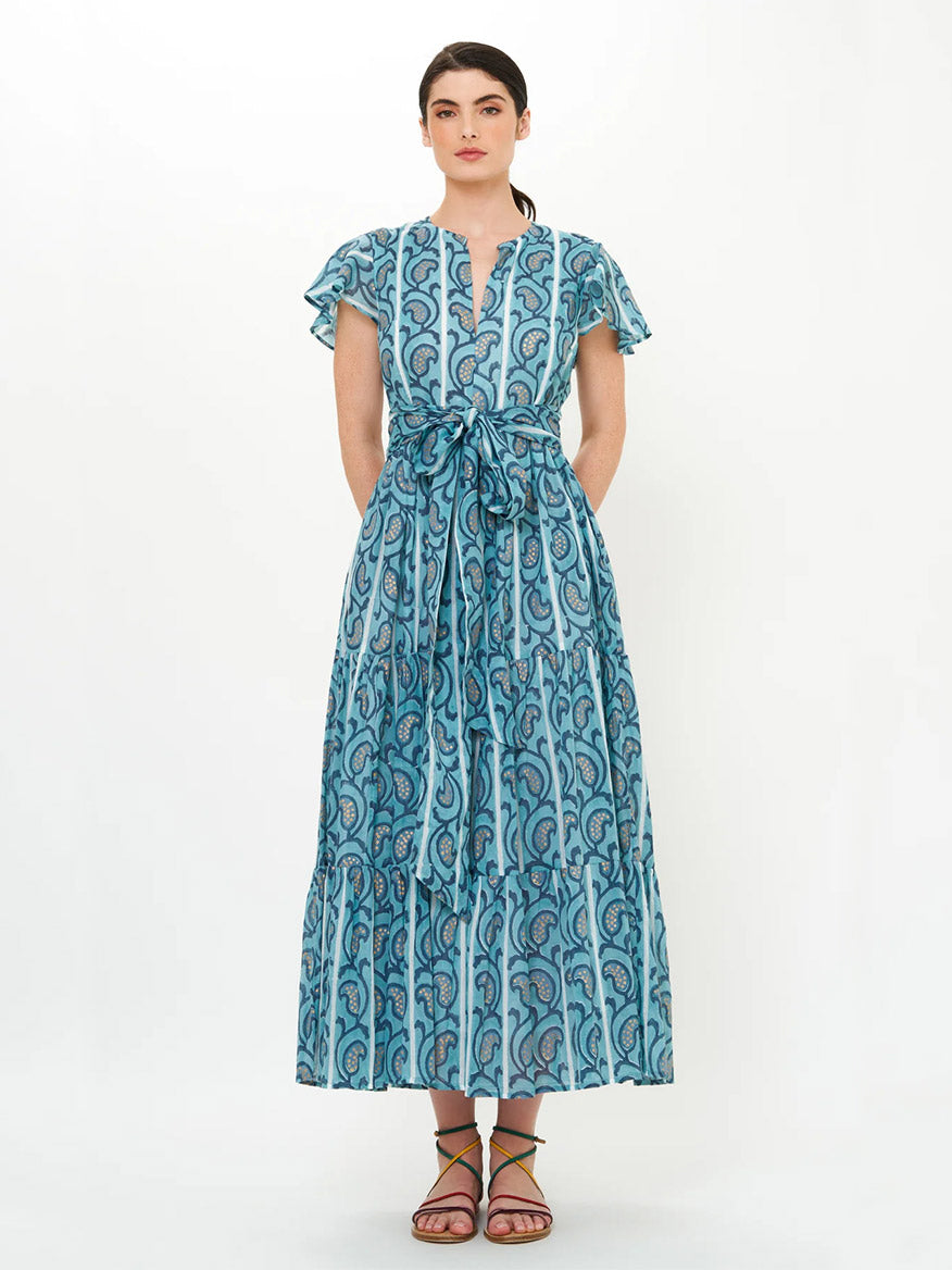 A woman wears the Oliphant V-Neck Maxi Dress in Goa Blue, featuring hand block printing, short flutter sleeves, and a waist tie, paired with flat sandals against a plain background.