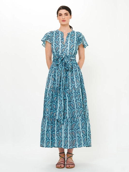 A woman wears the Oliphant V-Neck Maxi Dress in Goa Blue, featuring hand block printing, short flutter sleeves, and a waist tie, paired with flat sandals against a plain background.