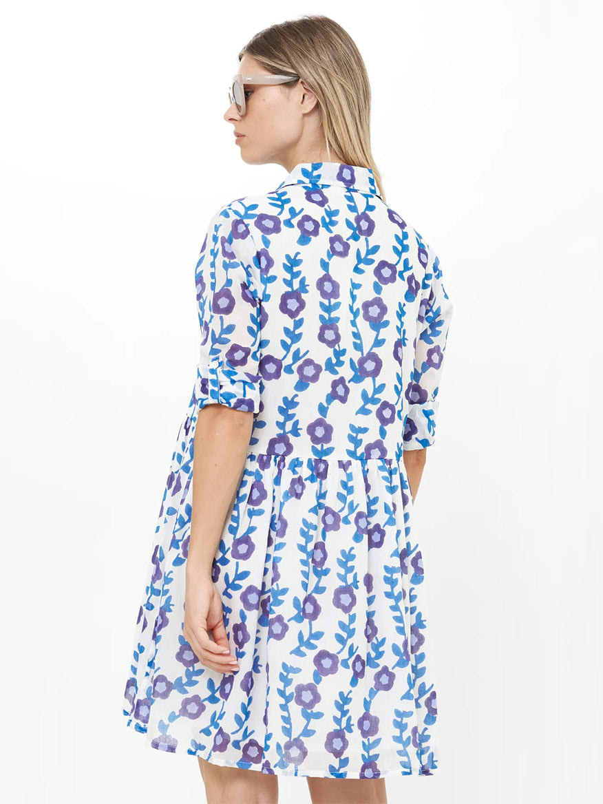 A woman with blonde hair radiates style and comfort, seen from the back in sunglasses and the Oliphant Long Sleeve Shirt Dress Mini in Toscana Blue, featuring blue and purple floral patterns.