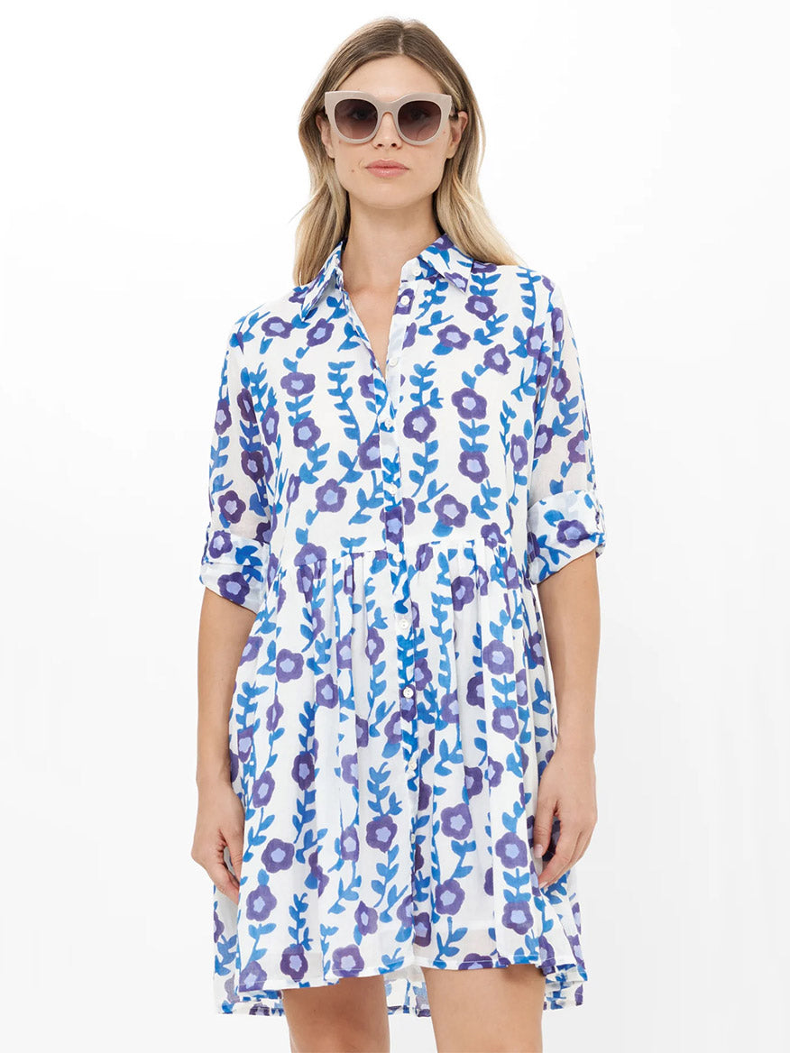 A woman models the Oliphant Long Sleeve Shirt Dress Mini in Toscana Blue, featuring blue and purple floral patterns, sunglasses, and a chic white backdrop—blending effortless style with comfort.