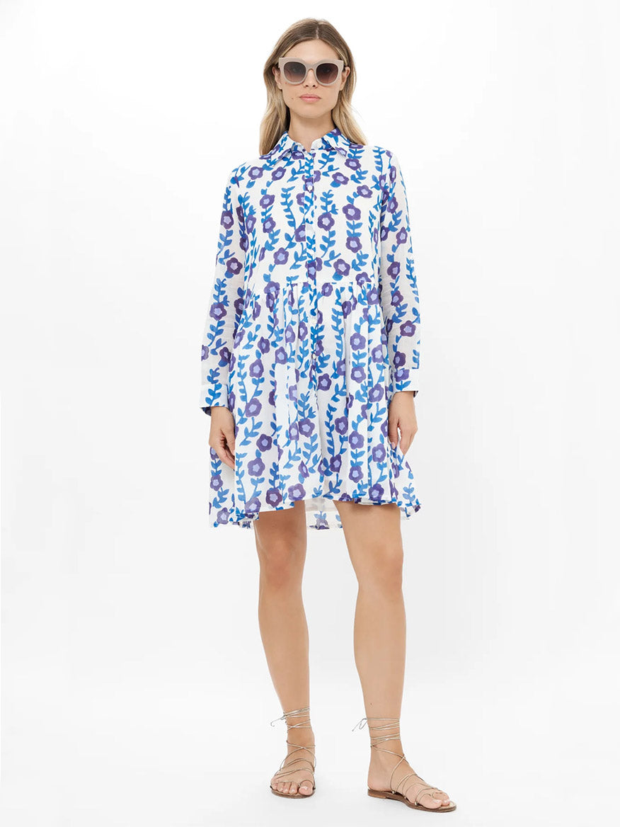 Woman wearing the Oliphant Long Sleeve Shirt Dress Mini in Toscana Blue with blue and purple floral patterns, styled for comfort, paired with sunglasses and flat sandals, standing against a plain white background.