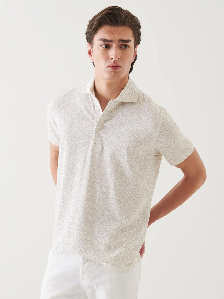 A young man stands against a plain background wearing the Patrick Assaraf Pima Cotton Stretch Printed Polo in Clay and white pants, with his hands behind his back.