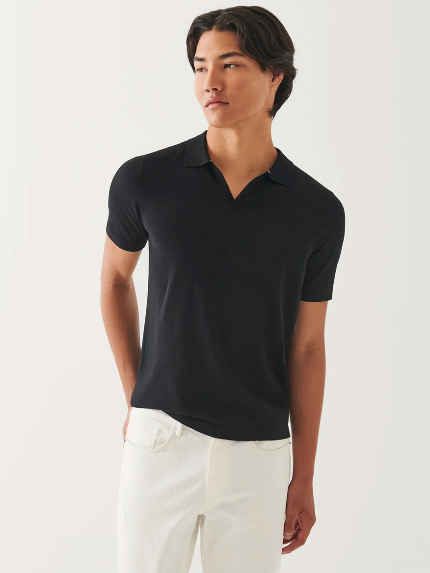 A person wears the Patrick Assaraf Cotton Cupro Open Polo in Black with white pants, standing against a plain light background and gazing to the side—showcasing versatile styling for any occasion.