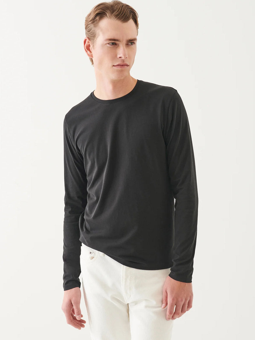 A person wears the Patrick Assaraf Pima Cotton Stretch Long Sleeve T-Shirt in Black with white pants, standing against a plain white background and looking slightly to the side.