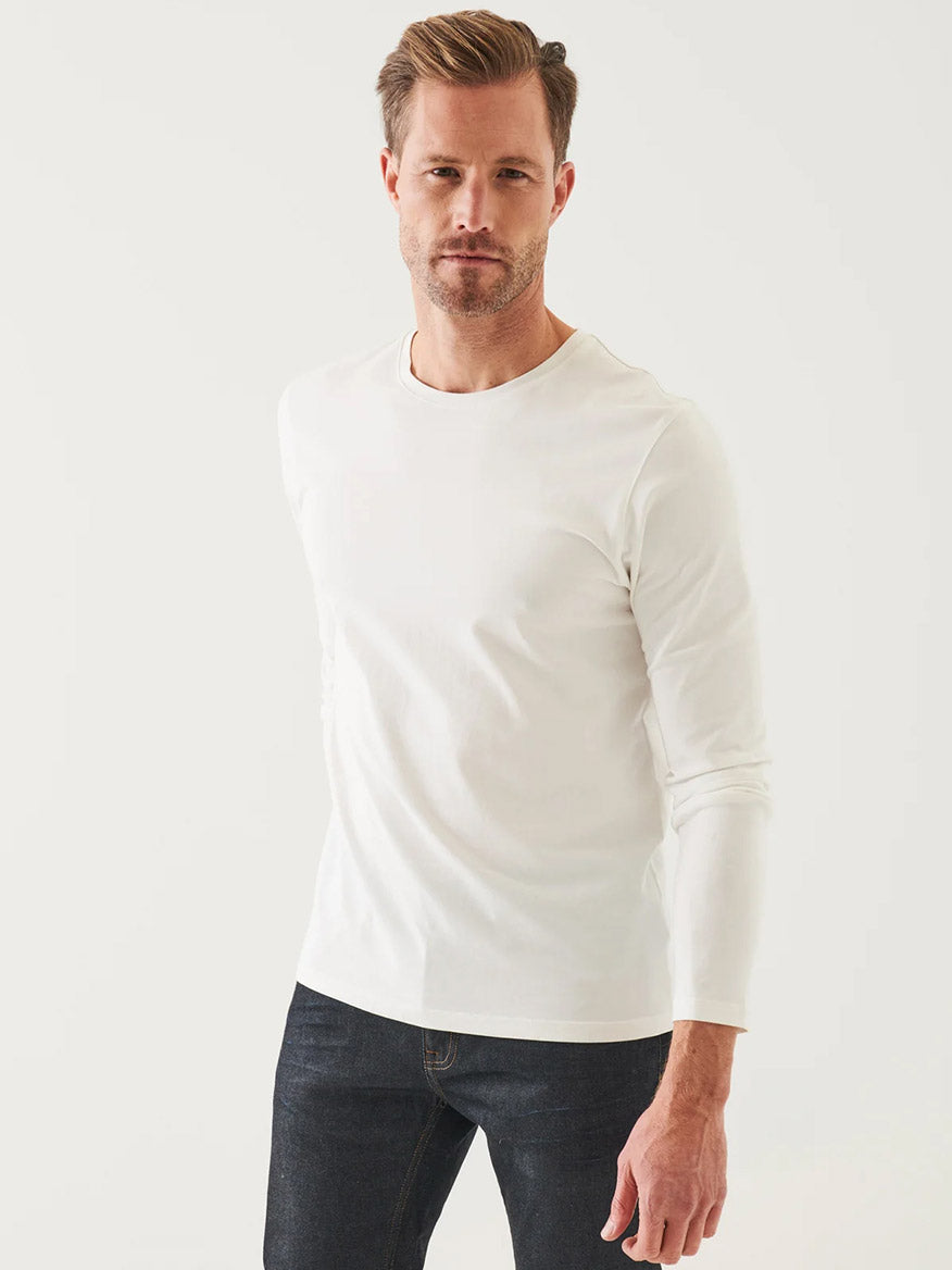 A man wearing a Patrick Assaraf Pima Cotton Stretch Long Sleeve T-Shirt in white and dark jeans stands against a light background, looking at the camera.