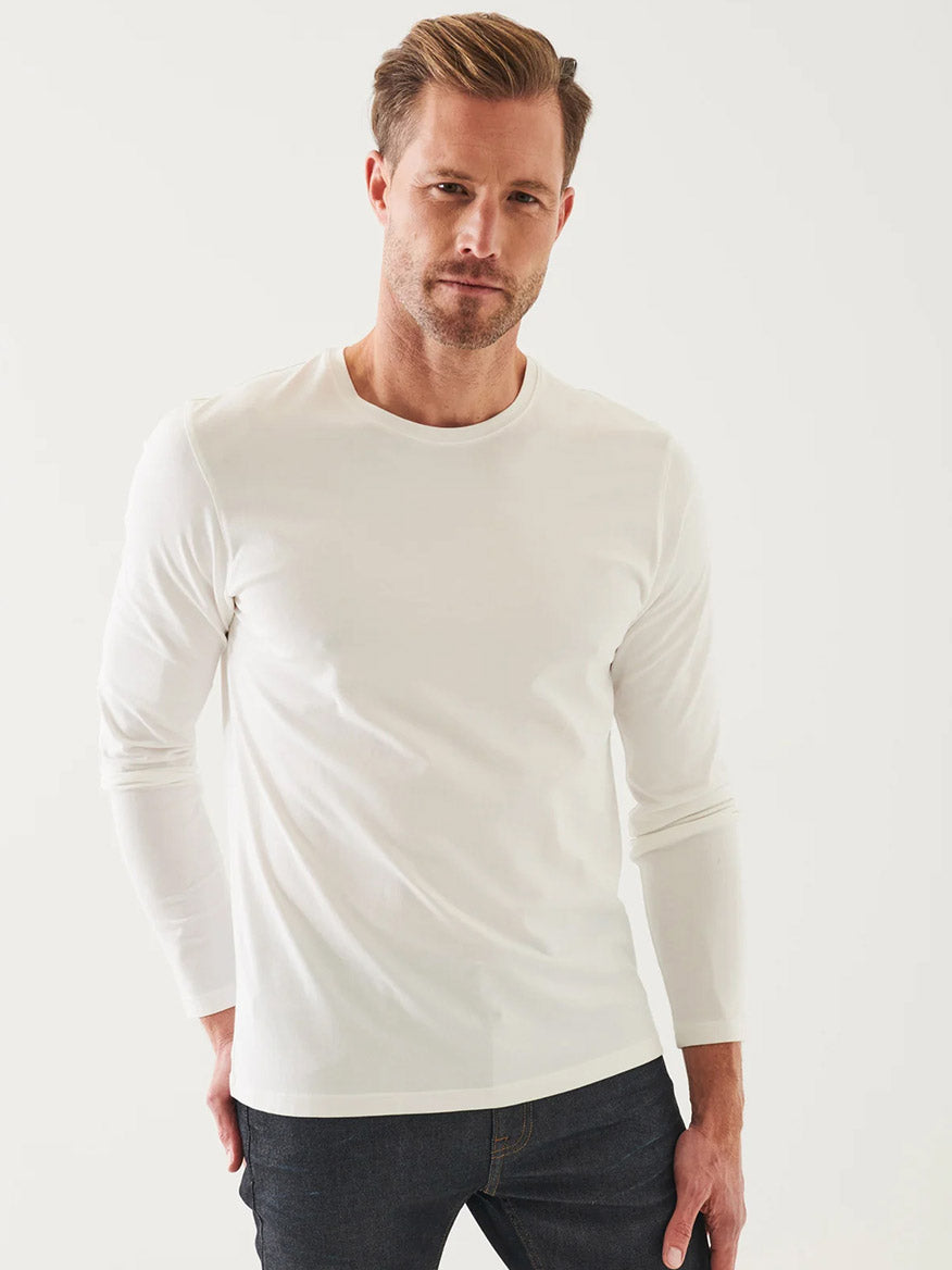 A man wearing a Patrick Assaraf Pima Cotton Stretch Long Sleeve T-Shirt in White and dark jeans stands against a plain light background, looking directly at the camera.