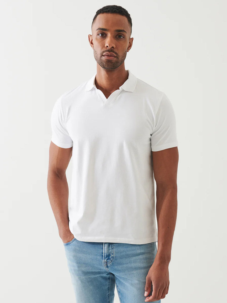 A man models the Patrick Assaraf Pima Cotton Stretch Open Polo in White with light blue jeans against a simple, light background, highlighting a versatile and timeless style.