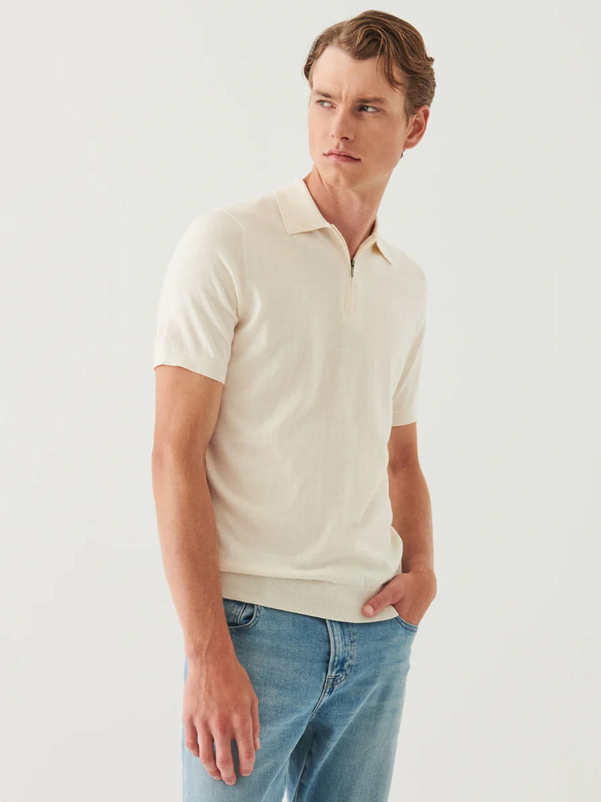 A man wearing the Patrick Assaraf Cotton Cashmere Quarter Zip Polo in Off White and blue jeans stands against a plain light background, looking to his left with his right hand in his pocket.