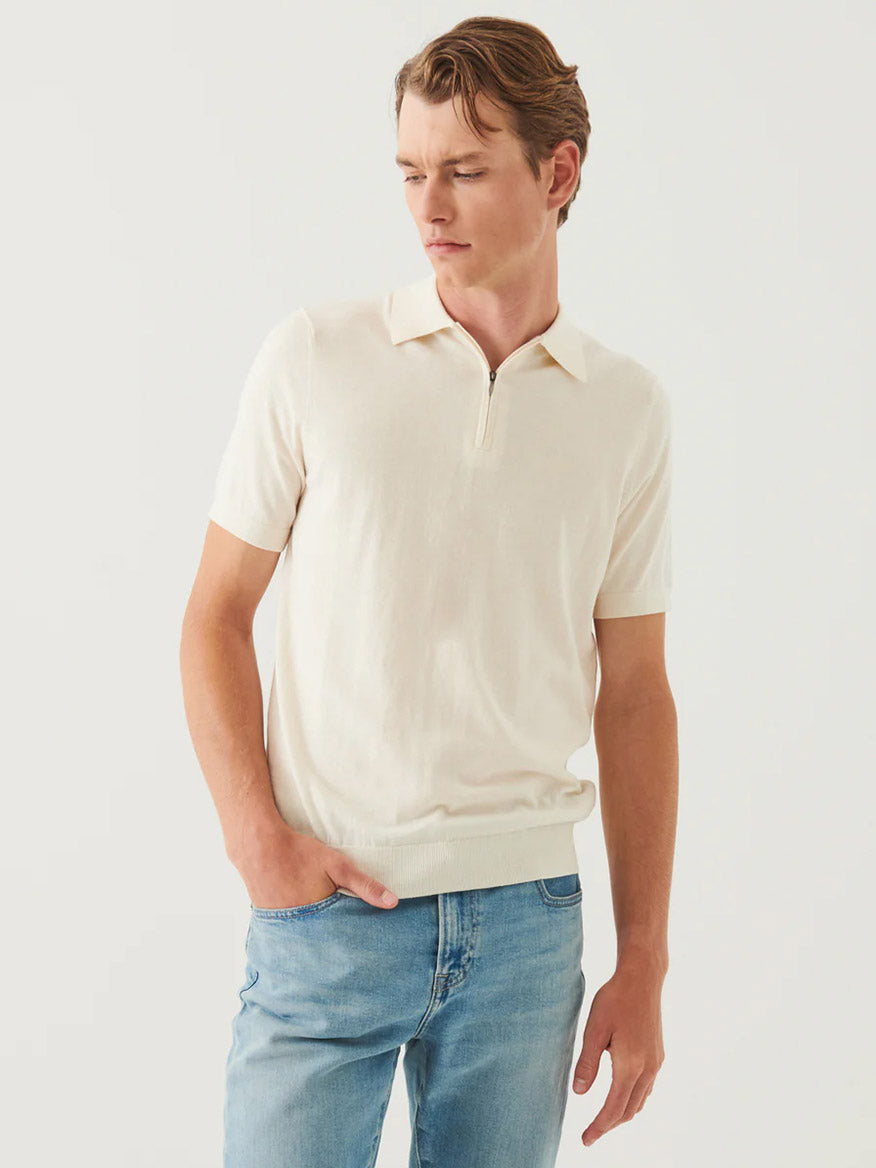 A man wearing a Patrick Assaraf Cotton Cashmere Quarter Zip Polo in Off White and blue jeans stands with one hand in his pocket, looking down to the side against a plain background.