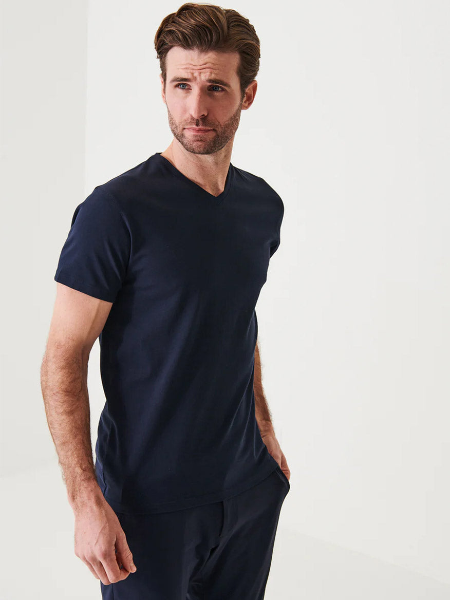 A man with brown hair and a trimmed beard wears the Patrick Assaraf Pima Cotton Stretch V-Neck T-Shirt in Midnight with dark pants, standing against a white background—showcasing timeless style.
