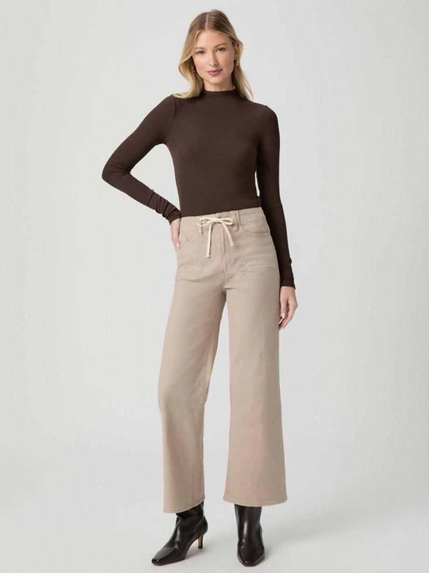 A woman stands facing forward in a fitted long-sleeve brown top and Paige Ari Wide Leg Pant in Vintage Utopia, paired with black heeled ankle boots, against a plain light background.