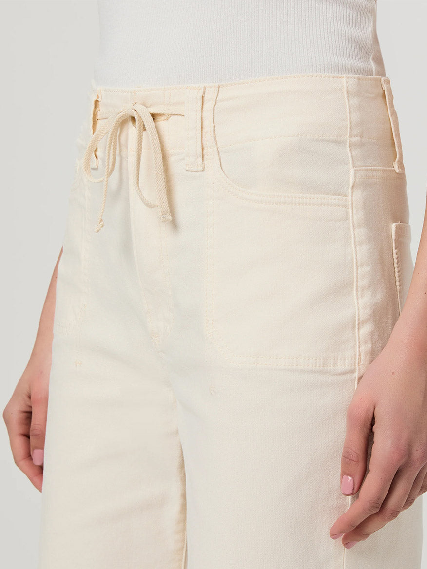 A person wearing the Paige Ari Wide Leg Pant in Quartz Sand, featuring a cream color with a high-rise drawstring waist and front pockets, paired with a white top.