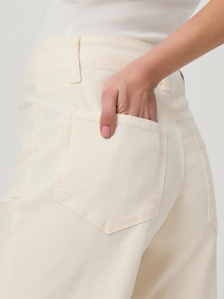 A person in a white top is casually resting a hand in the back pocket of Paige Ari Wide Leg Pant in Quartz Sand, which features cream-colored, super soft denim with wide legs.
