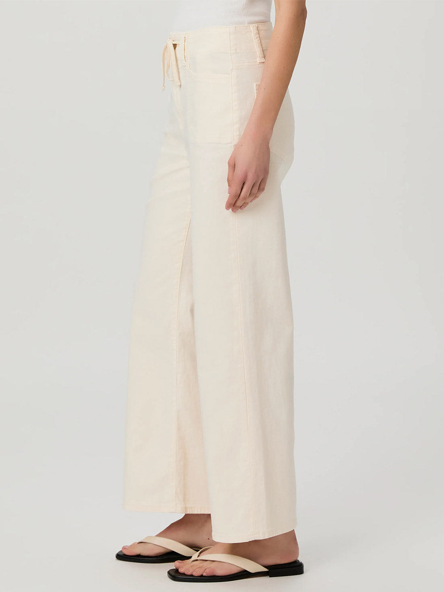 Wearing Paige Ari Wide Leg Pant in Quartz Sand, styled with sandals, an individual strikes a sideways pose against a plain backdrop, highlighting the pant's high-rise cut that exudes elegance.