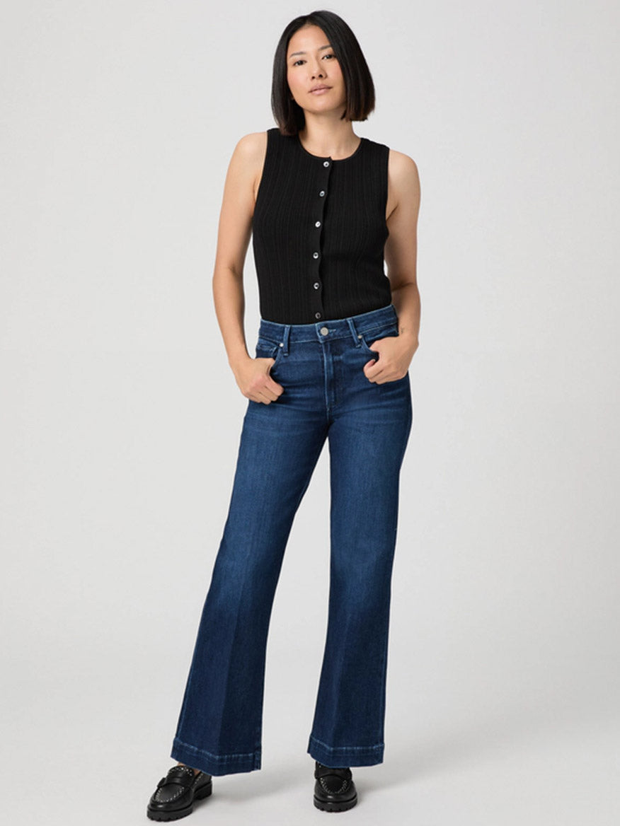 A woman stands with hands in her pockets, wearing a black sleeveless button-up top, Paige Leenah Petite Wide Leg Jean in Praline, and black shoes against a plain light background.