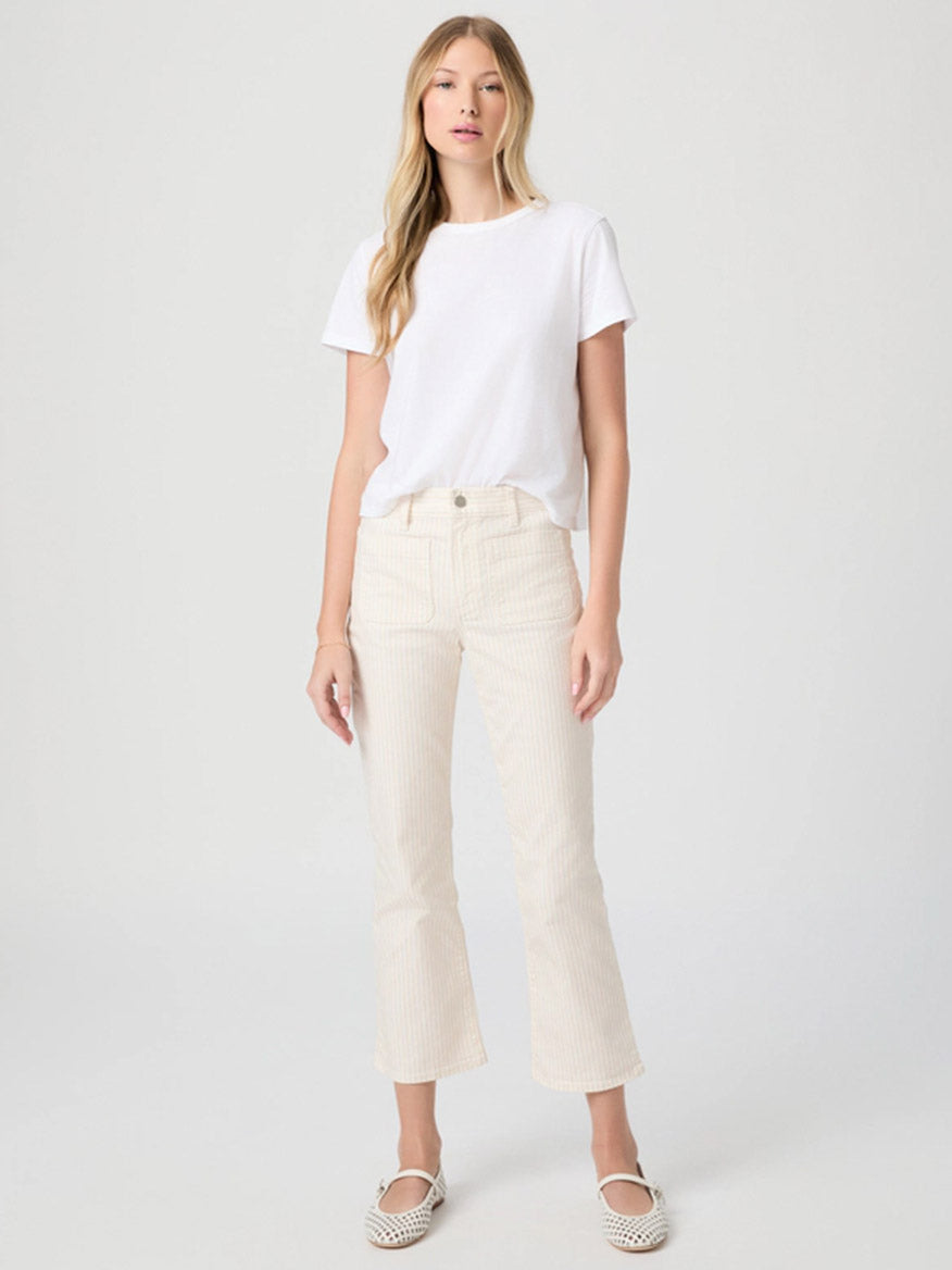 A woman stands against a plain background wearing a white t-shirt, slip-on shoes, and Paige Mason Crop Bootcut Jean in Tonal Stripe.