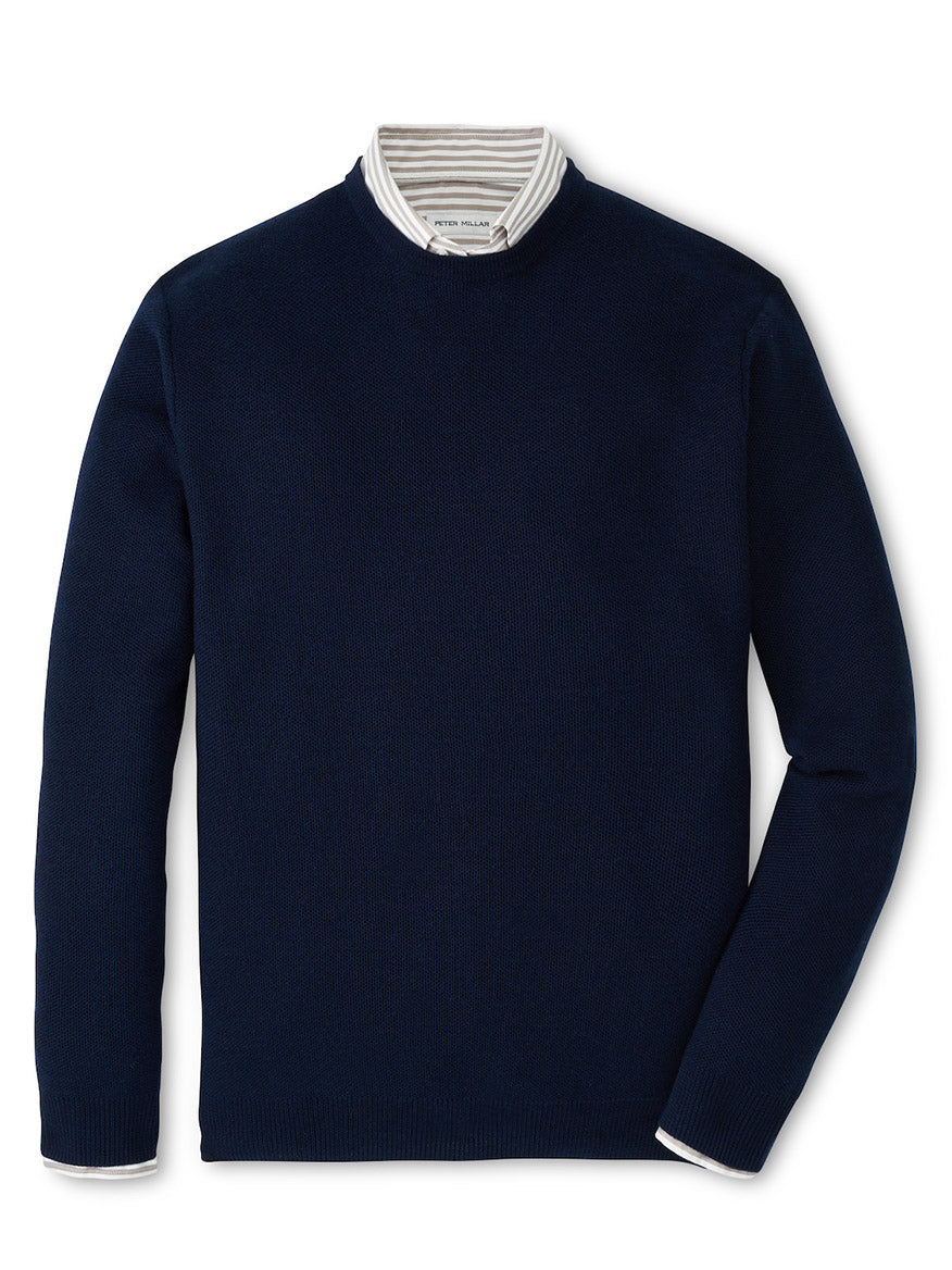 The Peter Millar Autumn Crest Honeycomb Crewneck Sweater in Navy is shown layered over a white collared shirt with gray stripes and displayed on a flat surface.
