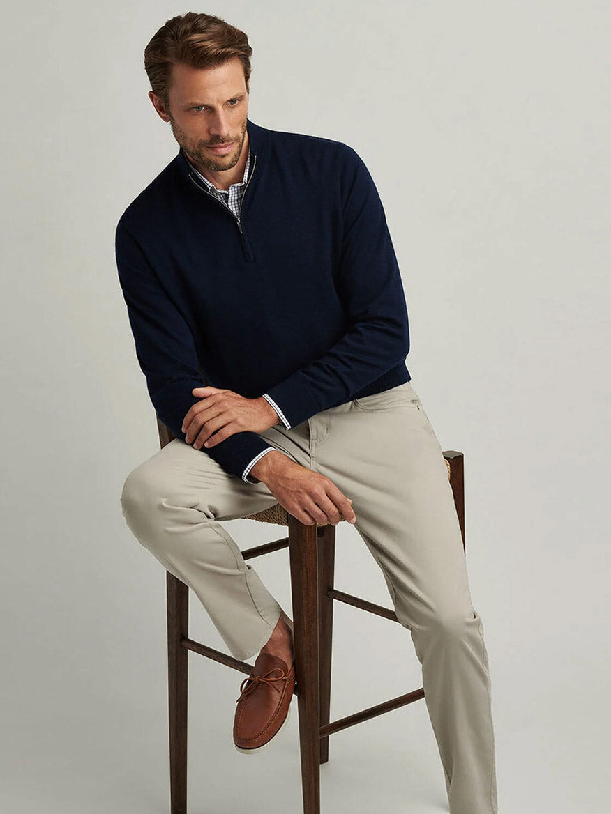 A man sits on a wooden stool against a plain light background wearing the Peter Millar Autumn Crest Quarter-Zip in Navy, paired with light khaki pants, brown shoes, and a checked shirt.