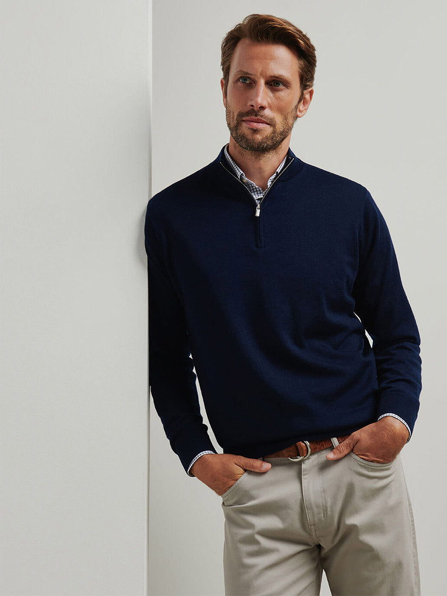 A man with light brown hair and a beard stands against a white wall, wearing the Peter Millar Autumn Crest Quarter-Zip in Navy over a collared shirt and light-colored pants.