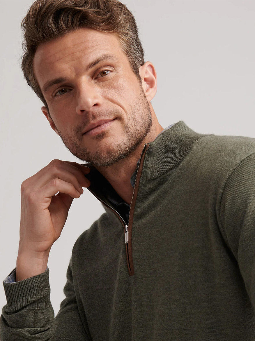 A man wearing the Peter Millar Autumn Crest Suede Trim Quarter-Zip in Basil looks at the camera while adjusting the zipper with his left hand.