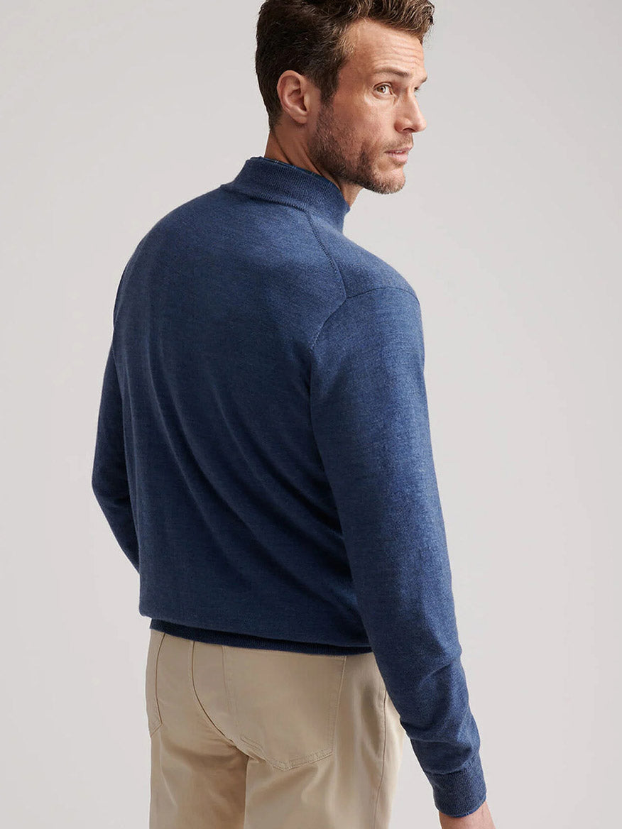 A man in the Peter Millar Autumn Crest Suede Trim Quarter-Zip in Blue Haze and beige pants stands facing away, glancing over his shoulder against a plain light background.