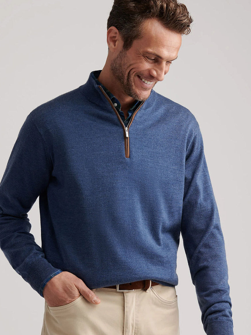 A man wears the Peter Millar Autumn Crest Suede Trim Quarter-Zip in Blue Haze over a collared shirt and beige pants, standing with one hand in his pocket as he looks down and smiles.