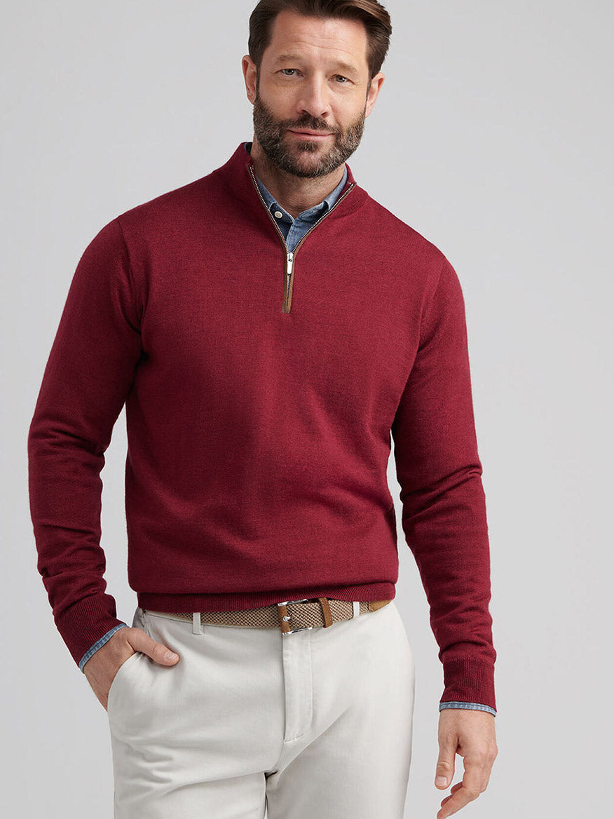 A bearded man wears the Peter Millar Autumn Crest Suede Trim Quarter-Zip in Brick Red over a blue collared shirt and beige pants, standing with one hand in his pocket.