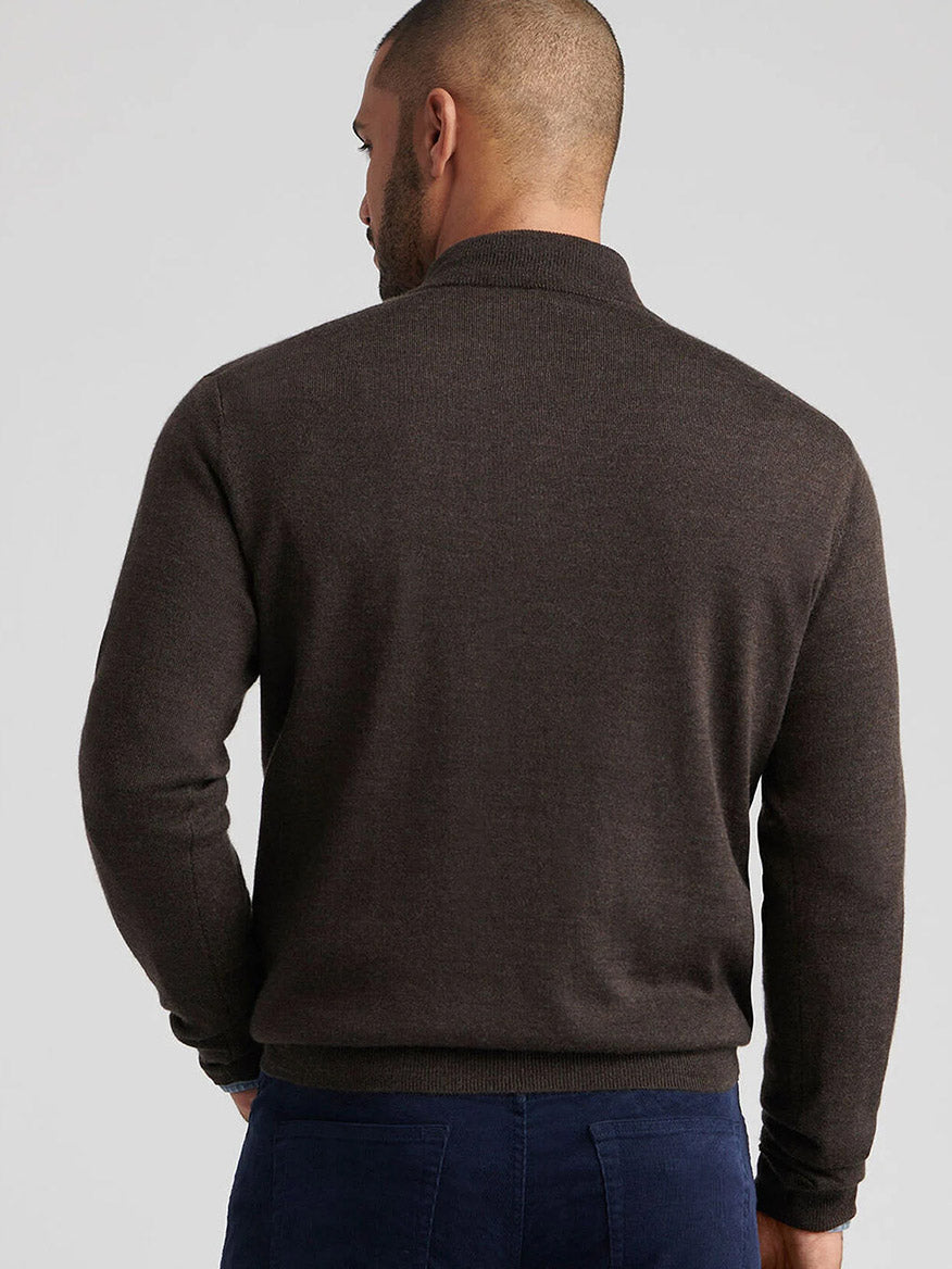 A man stands facing away from the camera, wearing the Peter Millar Autumn Crest Suede Trim Quarter-Zip in French Press and navy blue pants, showcasing a classic fit against a neutral background.