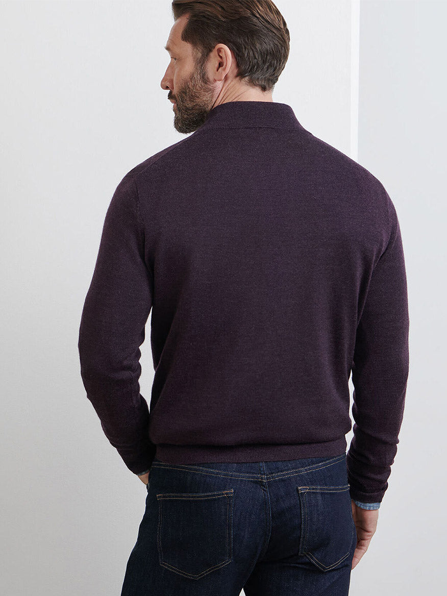 A man stands with his back to the camera, wearing the Peter Millar Autumn Crest Suede Trim Quarter-Zip in Midnight Grape and blue jeans against a plain light-colored wall.