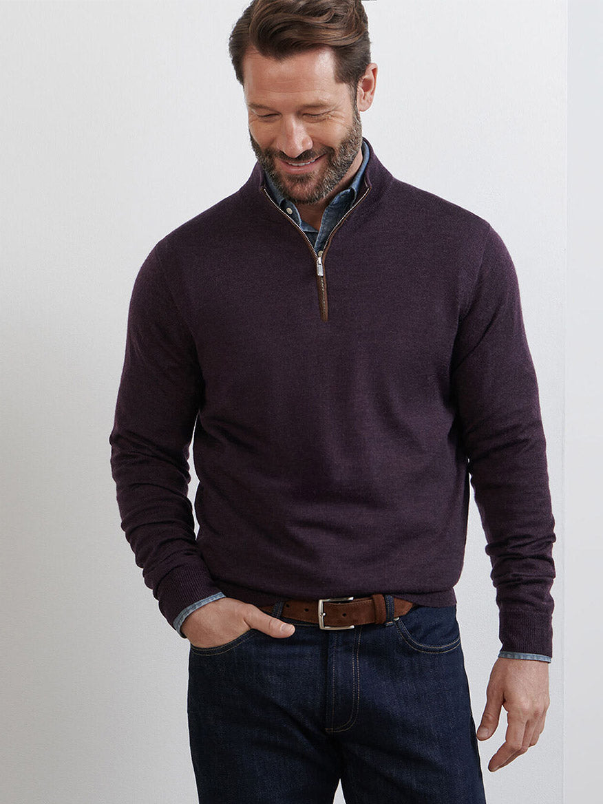 A man wears the Peter Millar Autumn Crest Suede Trim Quarter-Zip in Midnight Grape over a button-down shirt and jeans, standing against a plain light-colored wall while looking down and smiling.