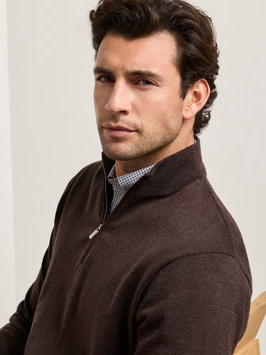 A man with short brown hair is seated and looking slightly to the side, wearing a collared shirt beneath the Peter Millar Autumn Crest Suede Trim Quarter-Zip in French Press (a classic-fit, dark brown Merino sweater).