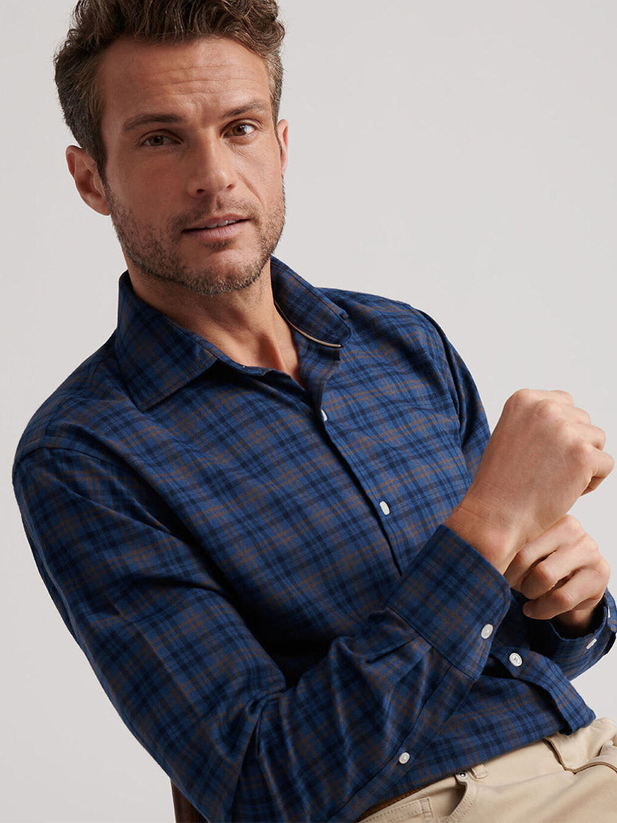 A man adjusts his sleeve while seated, wearing the Peter Millar Bar Harbor Performance Flannel Sport Shirt in Blue Haze, crafted from lightweight performance fibers, and looks toward the camera.