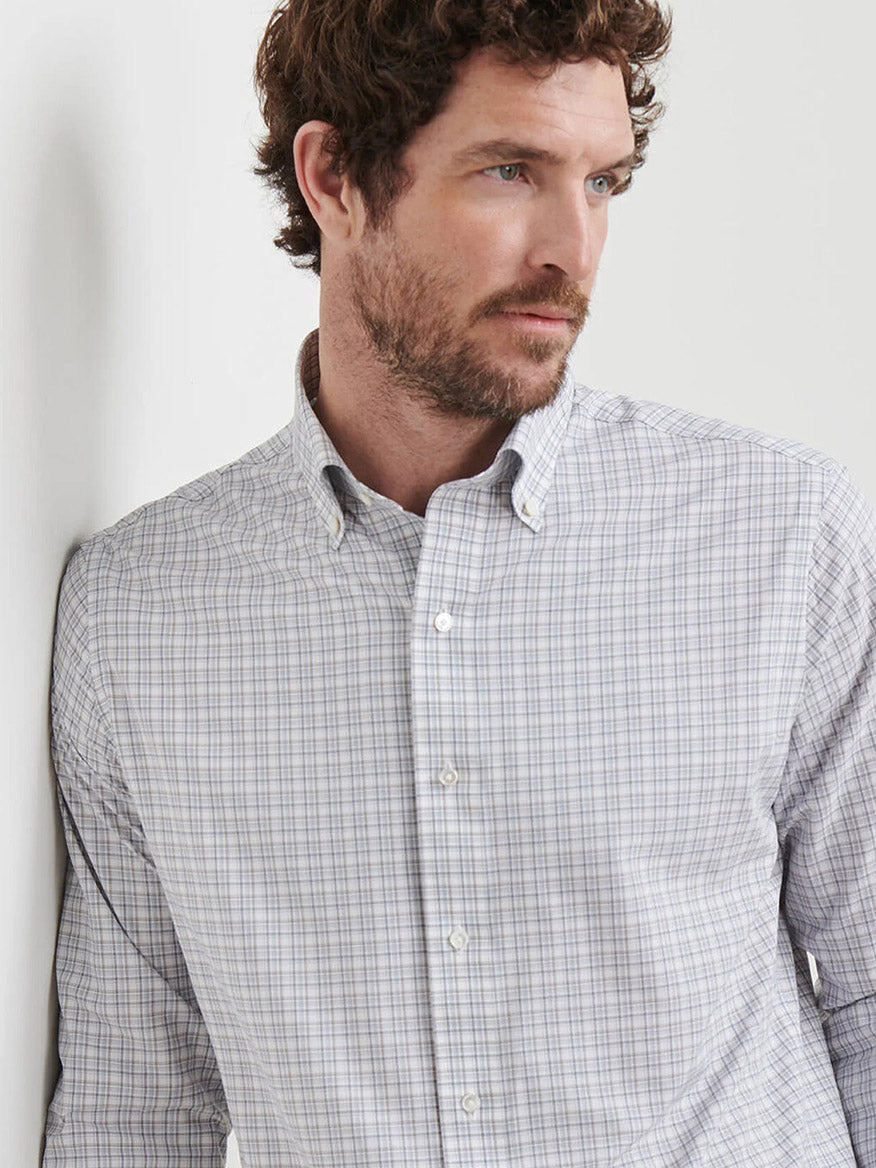A man with curly brown hair and a beard wears the Peter Millar Carter Performance Poplin Sport Shirt in Nebula Blue, looking to the side while standing against a white wall.