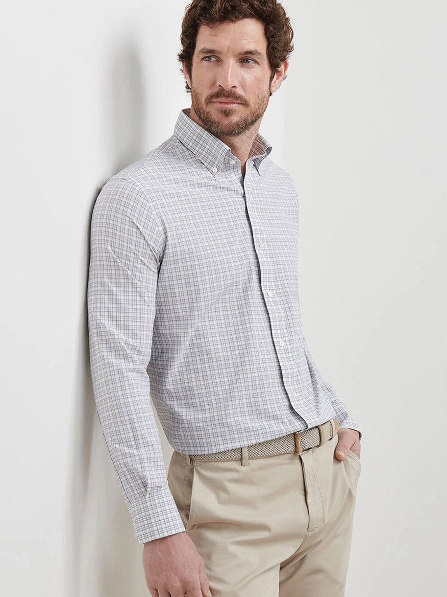 A man stands against a white wall with one hand in his pocket, wearing the Peter Millar Carter Performance Poplin Sport Shirt in Nebula Blue and beige pants.