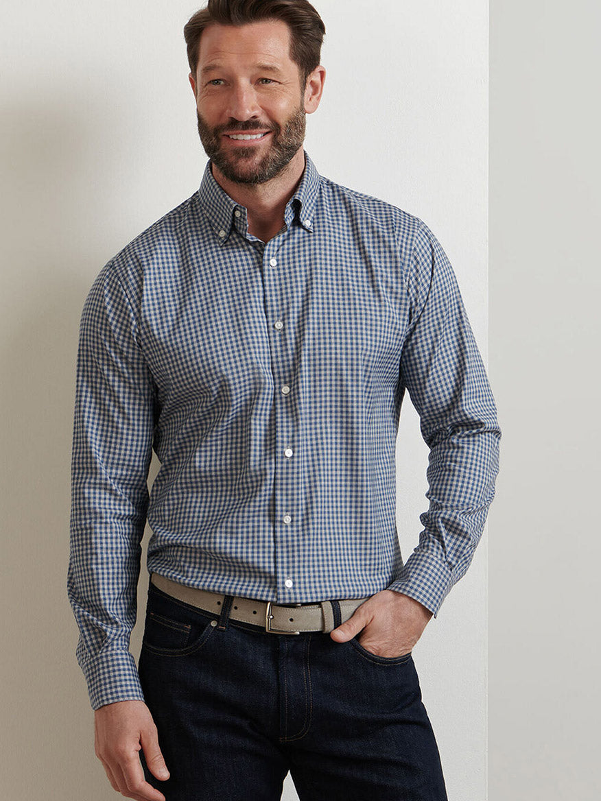 A man stands against a white wall, smiling with his left hand in his pocket, wearing the Peter Millar Conway Crown Lite Cotton-Stretch Sport Shirt in Gale Grey, dark jeans, and a light-colored belt.