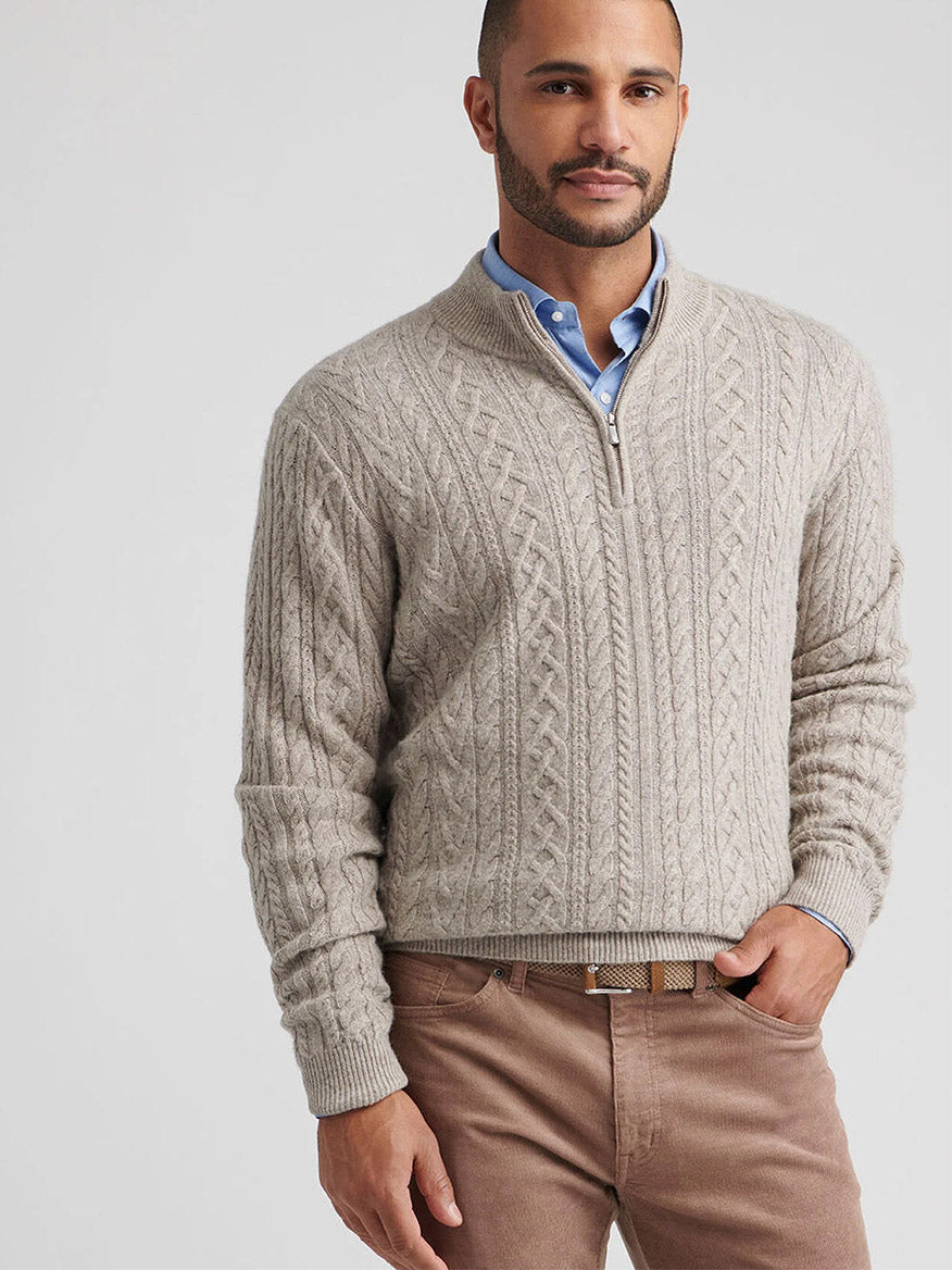 A man wears the Peter Millar Crescent Ridge Cable Quarter-Zip Sweater in Light Elm over a blue collared shirt with light brown pants, standing against a plain light background.