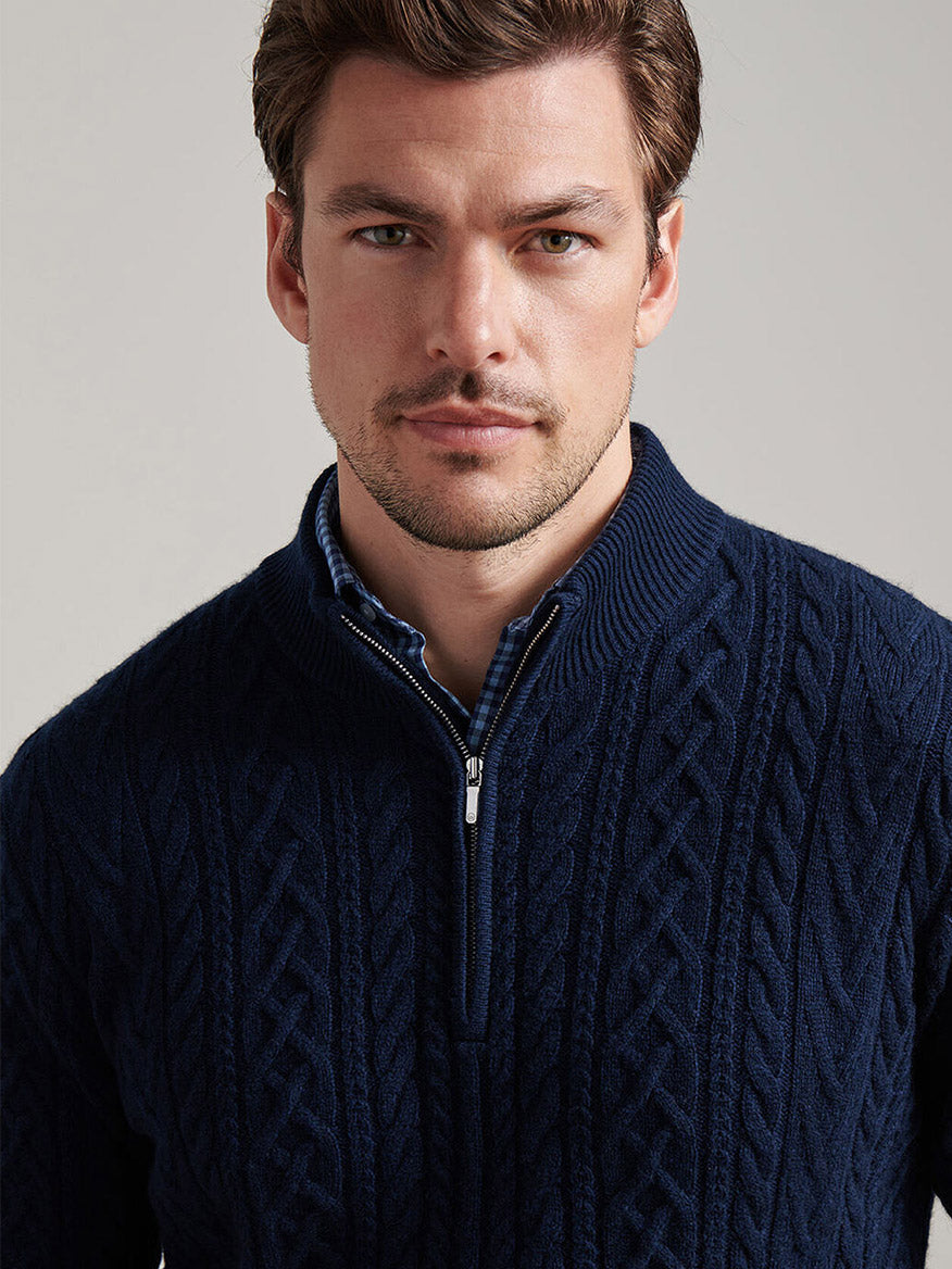A man with short brown hair and light stubble wears the Peter Millar Crescent Ridge Cable Quarter-Zip Sweater in Navy over a blue checkered shirt, looking directly at the camera.